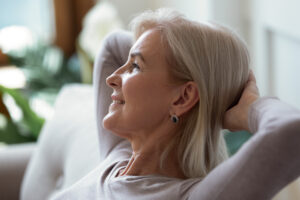 Older 60s woman put hands behind head resting seated indoors enjoy summer day looking out window close up pretty face side view, no stress, free time, serene calm person, keep calm, take break concept