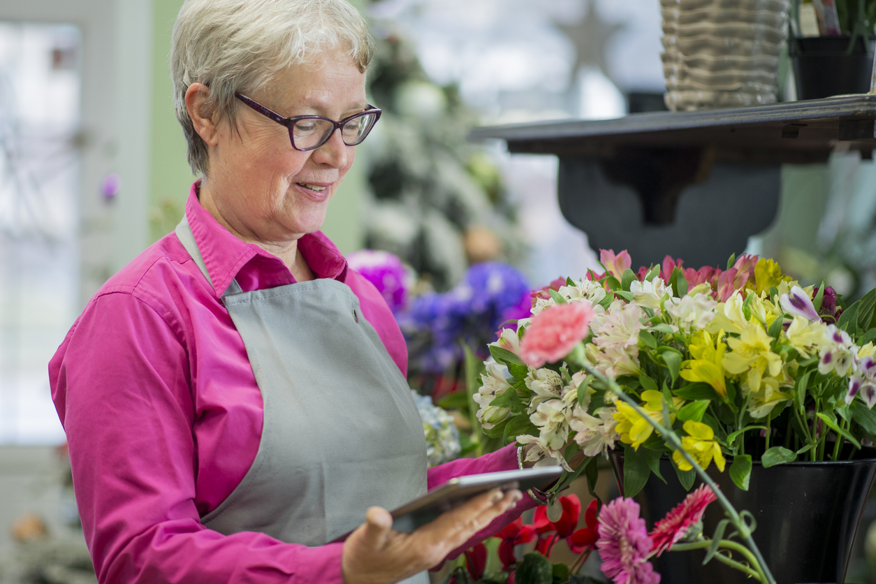A senior woman is the owner of a small flower shop. Here she takes inventory and inspects her products with a tablet computer.