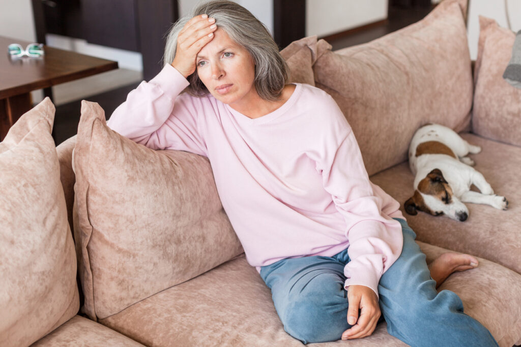Sad pensive middle age woman looking down depressed sitting on the sofa in the living room at home. Gray-haired female sitting on couch