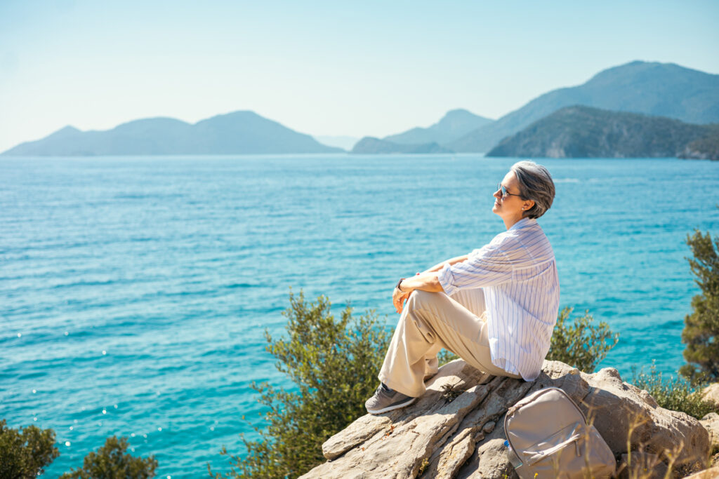 A senior woman with gray hair sits on a rocky cliff overlooking the bright blue sea, enjoying calm sunlight, fresh air, and a serene coastal moment during her travel getaway.